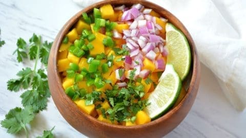 Wooden bowl with diced mango, jalapenos, onions and lime slices inside. A sprig of cilantro is off to the side.