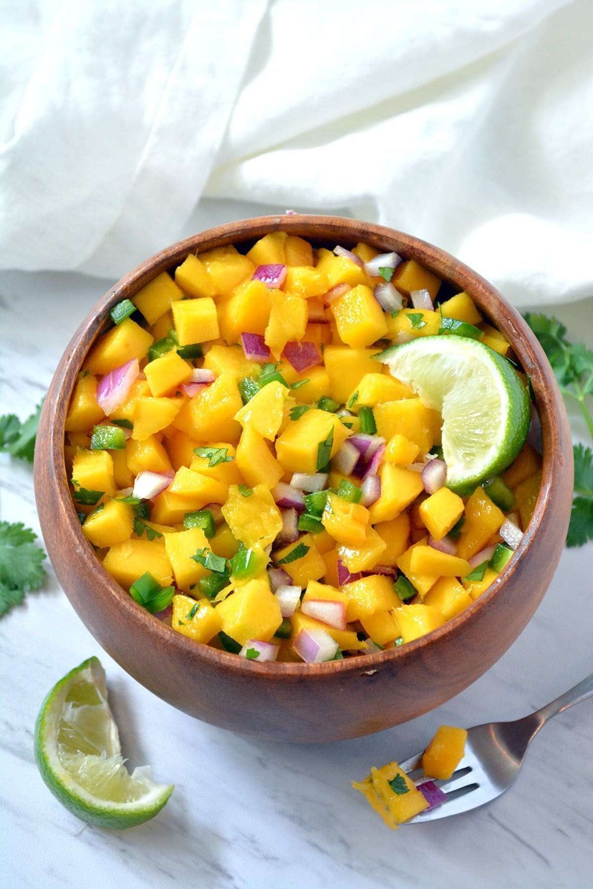 Wooden bowl with mango salsa and a wedge of lime inside.. A sprig of cilantro and lime wedge are off to the side.