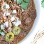 Close up of Birria in a white bowl topping with onions, pickled jalapenos and cilantro.