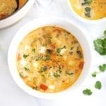 Two white bowls filled with Caldo de Queso Soup. Cilantro, black pepper, lime wedges and cubed cheese are next to the bowls.