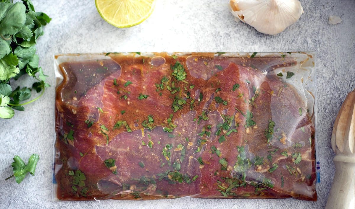 Close up of flank steak marinating in a ziploc bag next to lime, cilantro and garlic.