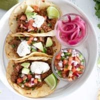 3 steak picado tacos on a white plate with topping options.