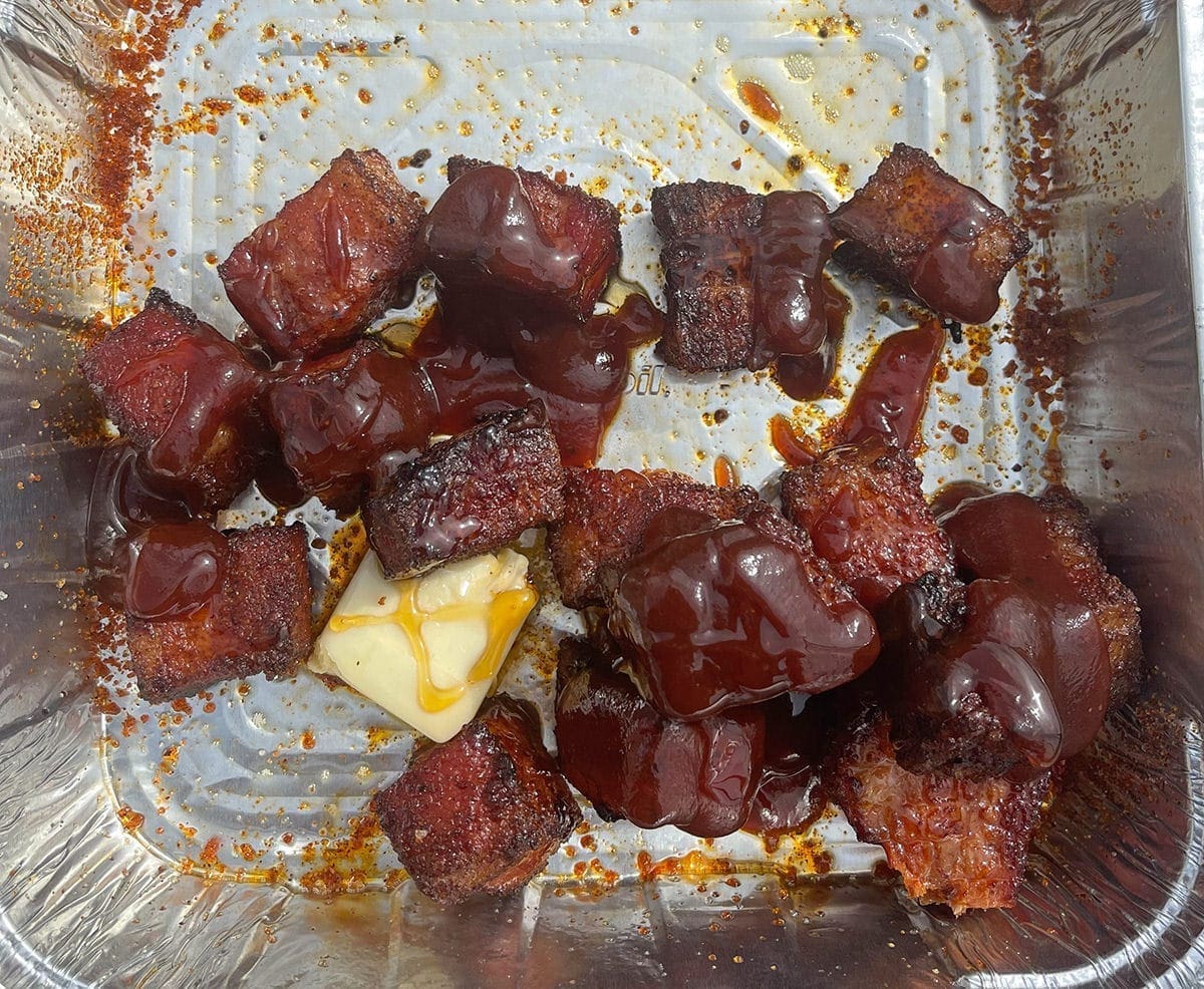 Pork Belly Burnt ends in a silver pan covered in BBQ sauce and butter.