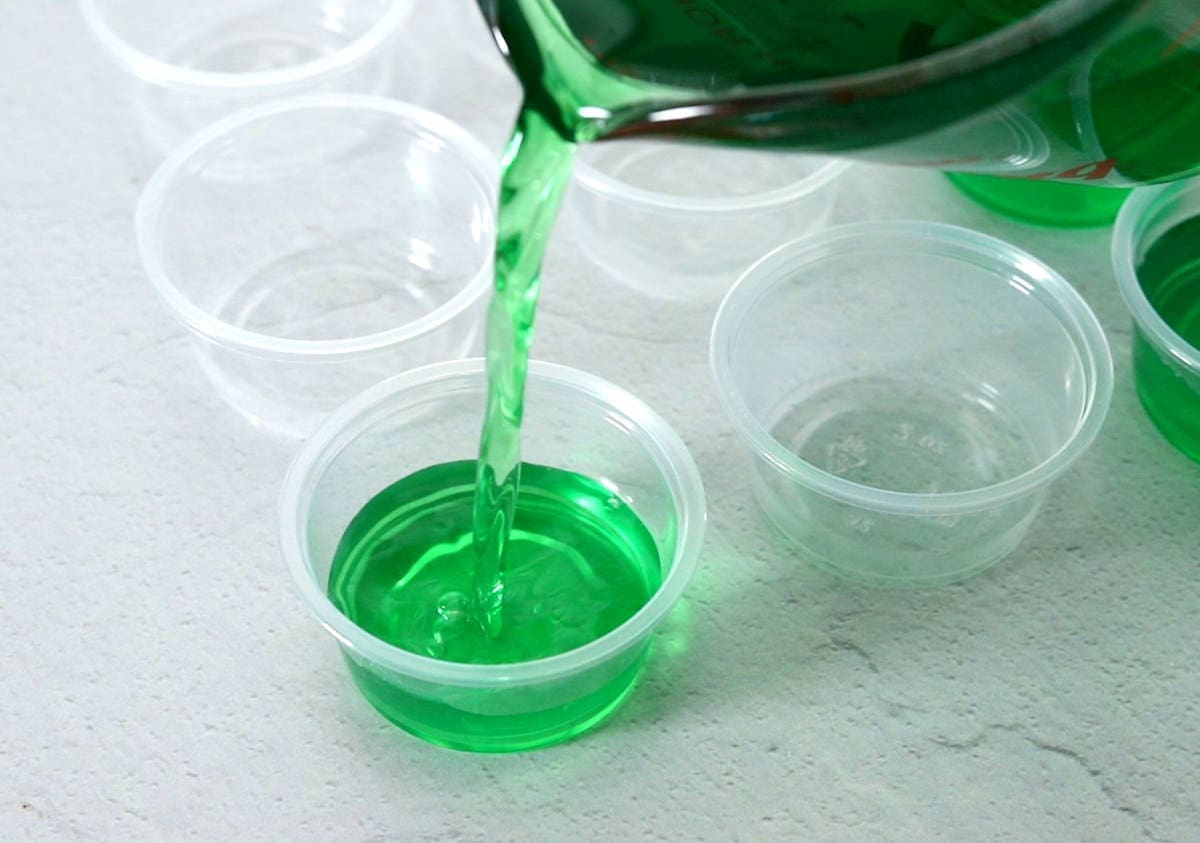 green jello being poured into a jello shot cup.