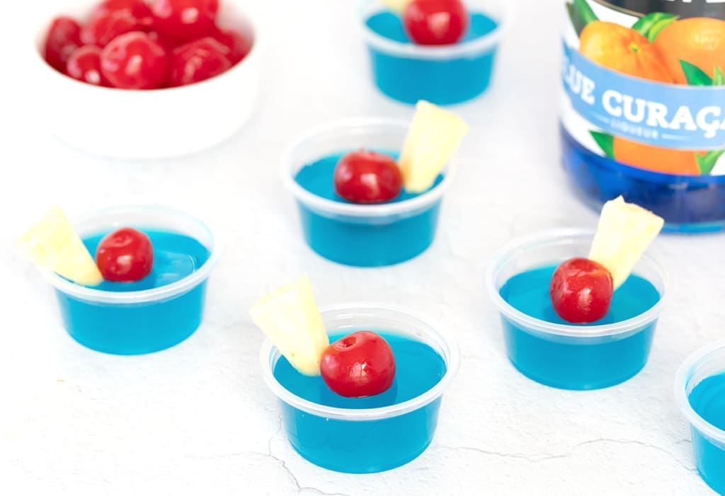 Multiple blue jello shots next to a bowl of cherries.