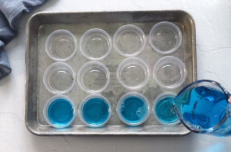 Baking sheet filled with plastic cups with jello being poured into them.