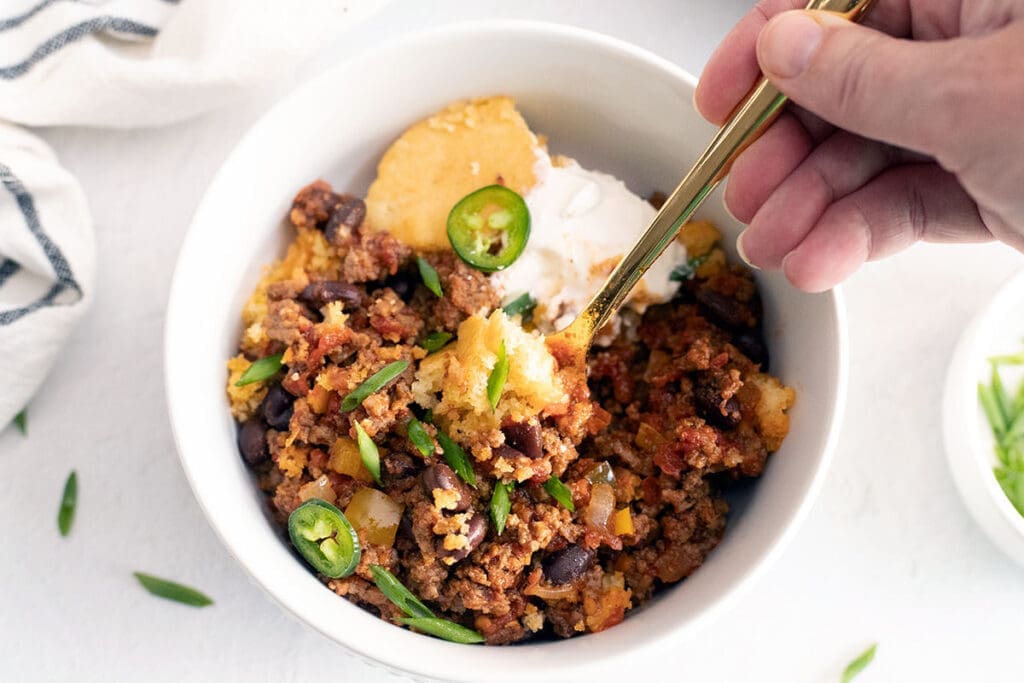 A hand holding a spoon digging into a bowl of cornbread with chili.