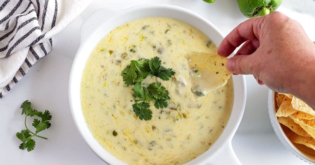 Hand holding a chip dipping into hatch green chile queso.