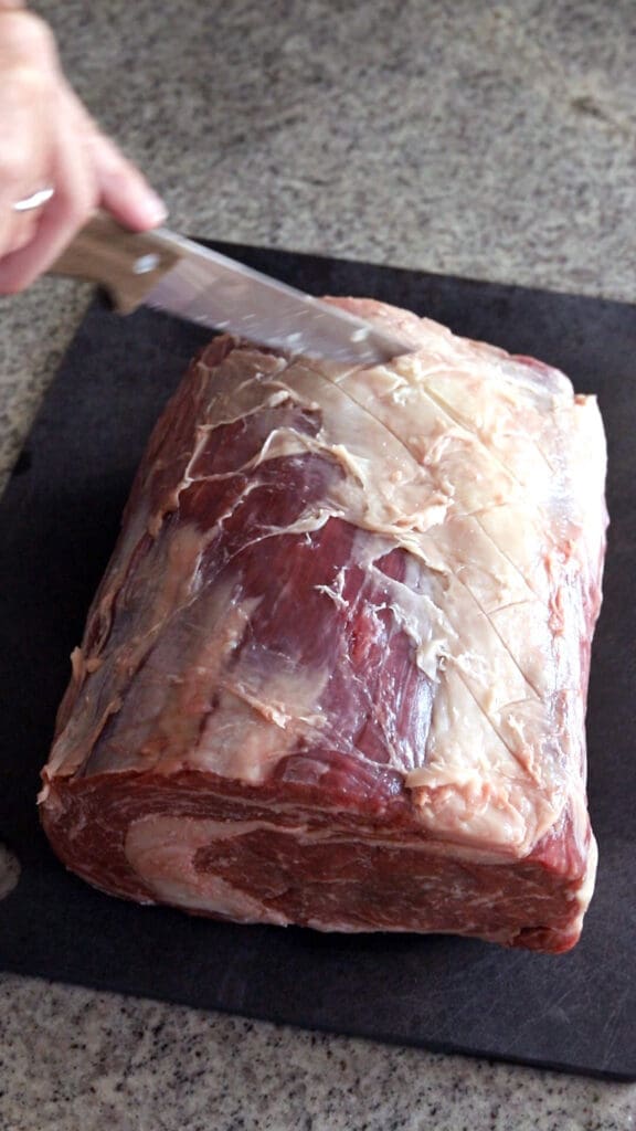 Hand trimming the fat off a prime rib roast on a black cutting board.