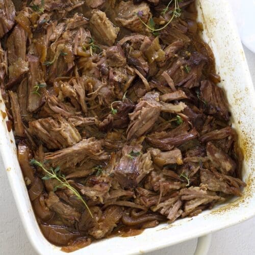 Chuck roast in the oven shredded in a white baking dish.