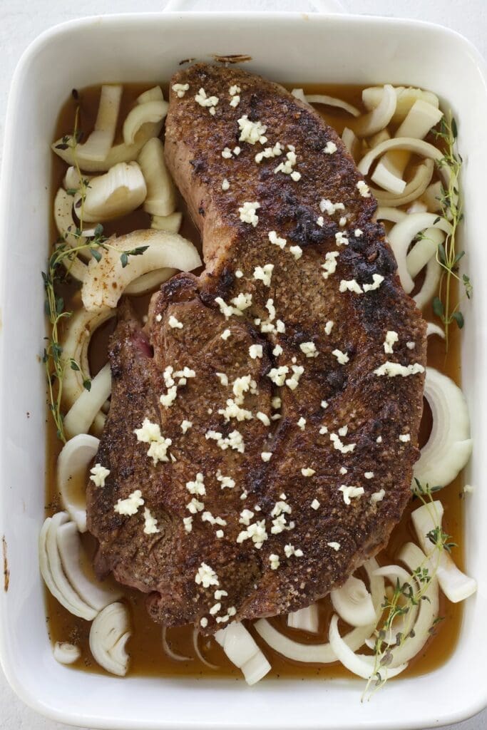 Beef covered in garlic with sliced onions in a baking pan.