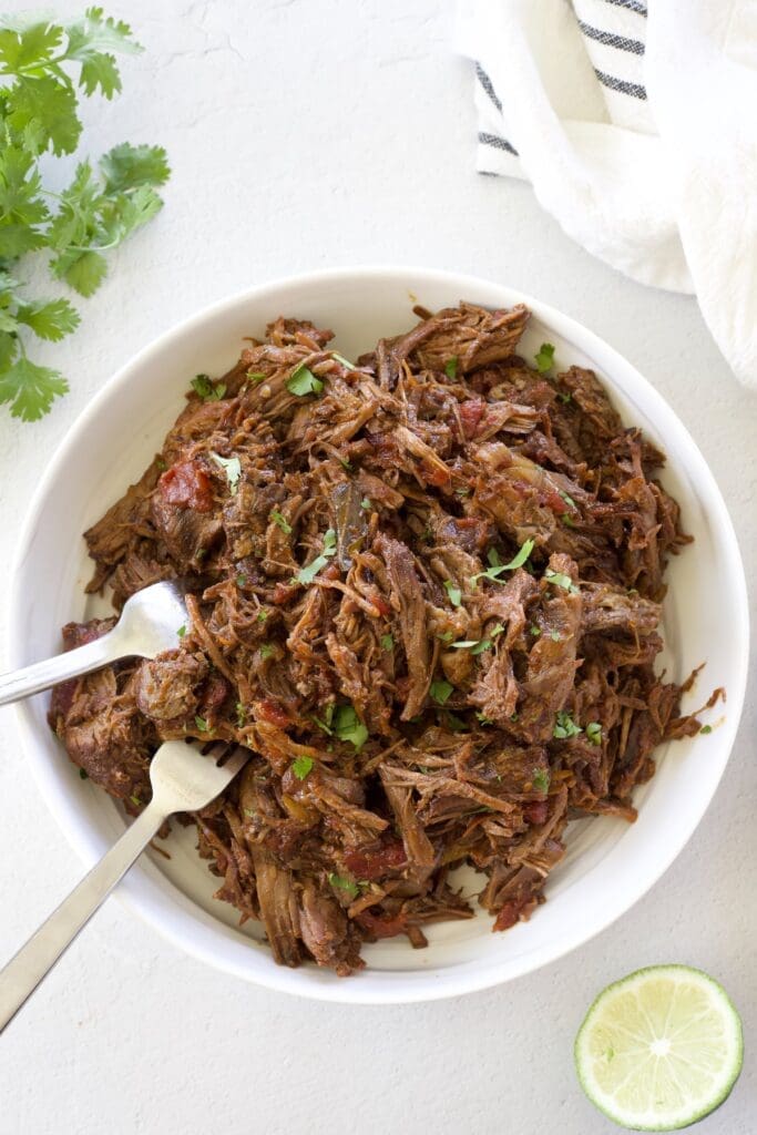 White bowl full of Mexican shredded beef and two forks.