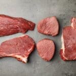 5 raw cuts of steak on a grey board for "cheapest cuts of beef" article.