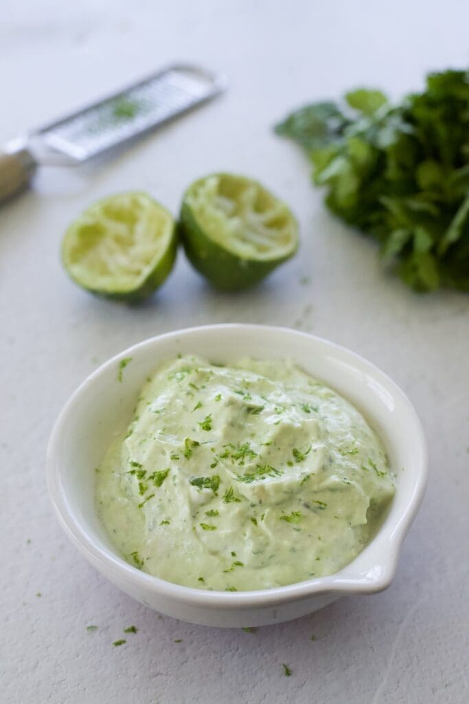 Bowl of green creamy sauce next to lime and cilantro.