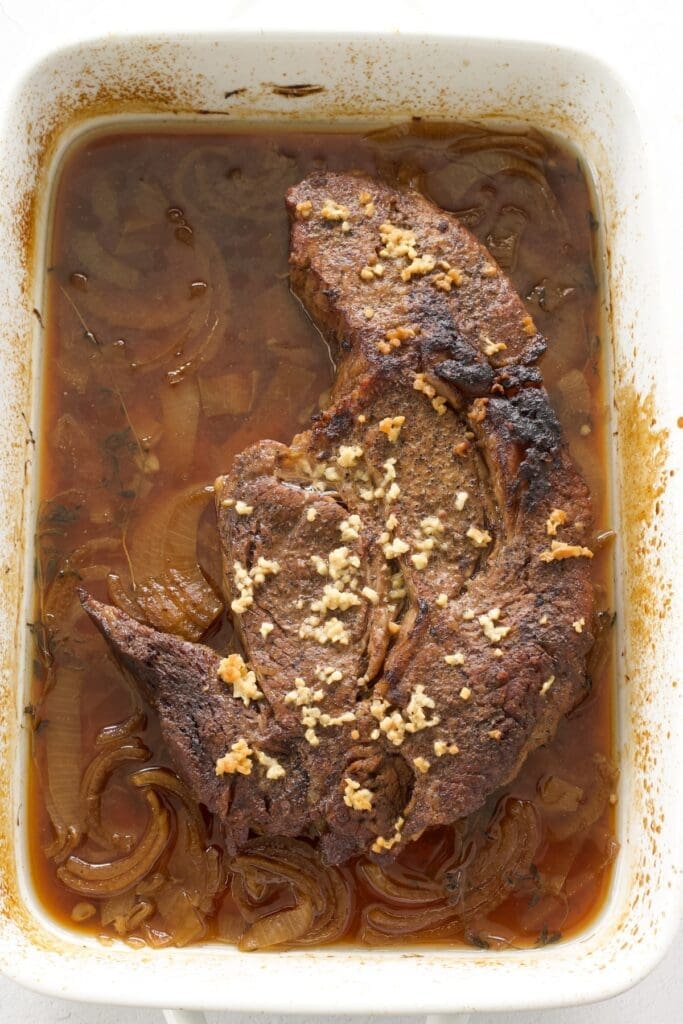 Cooked chuck roast in beef juice in a white baking pan.