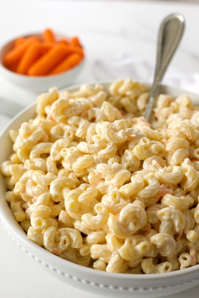 White bowl and spoon full of Hawaiian macaroni salad recipe with a bowl of carrots.