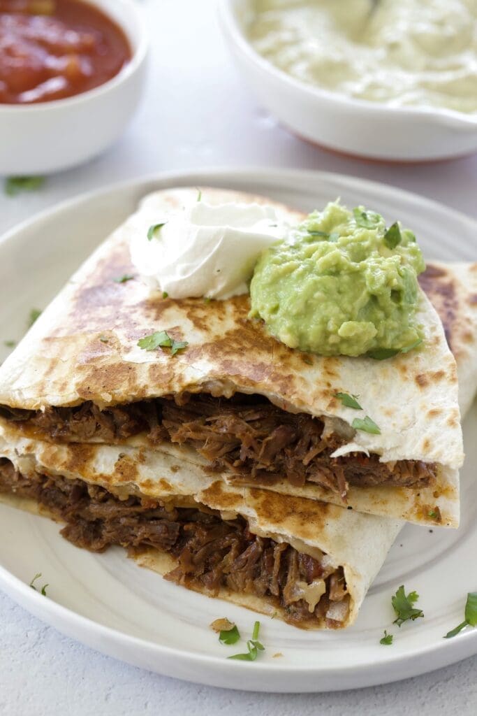 Shredded beef quesadillas on a white plate topped with sour cream and guacamole.