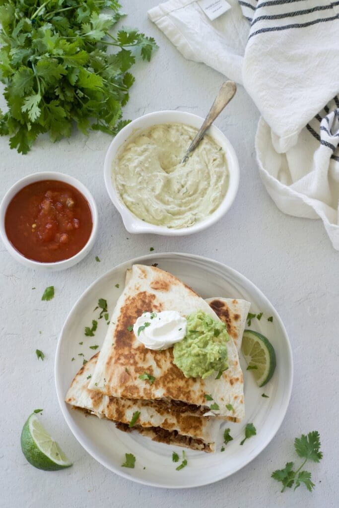 Shredded beef quesadillas on a white plate topped with sour cream and guacamole.